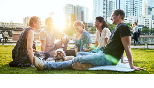 Drei Frauen, Zwei Männer und ein Hund sitzen im Gras und lachen zusammen. Im Hintergrund ist die Skyline von Frankfurt zu sehen.